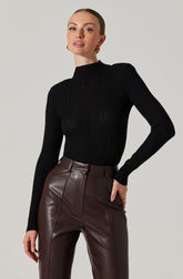 A woman is wearing a fitted black ribbed turtleneck sweater with long sleeves, paired with high-waisted brown leather pants that have front seam detailing.