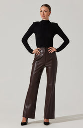 A woman is wearing a fitted black long-sleeve knit sweater with a high neckline. She pairs it with high-waisted, straight-leg dark brown leather pants and black pointed-toe heels. She has her hair slicked back and is accessorized with gold earrings and rings.