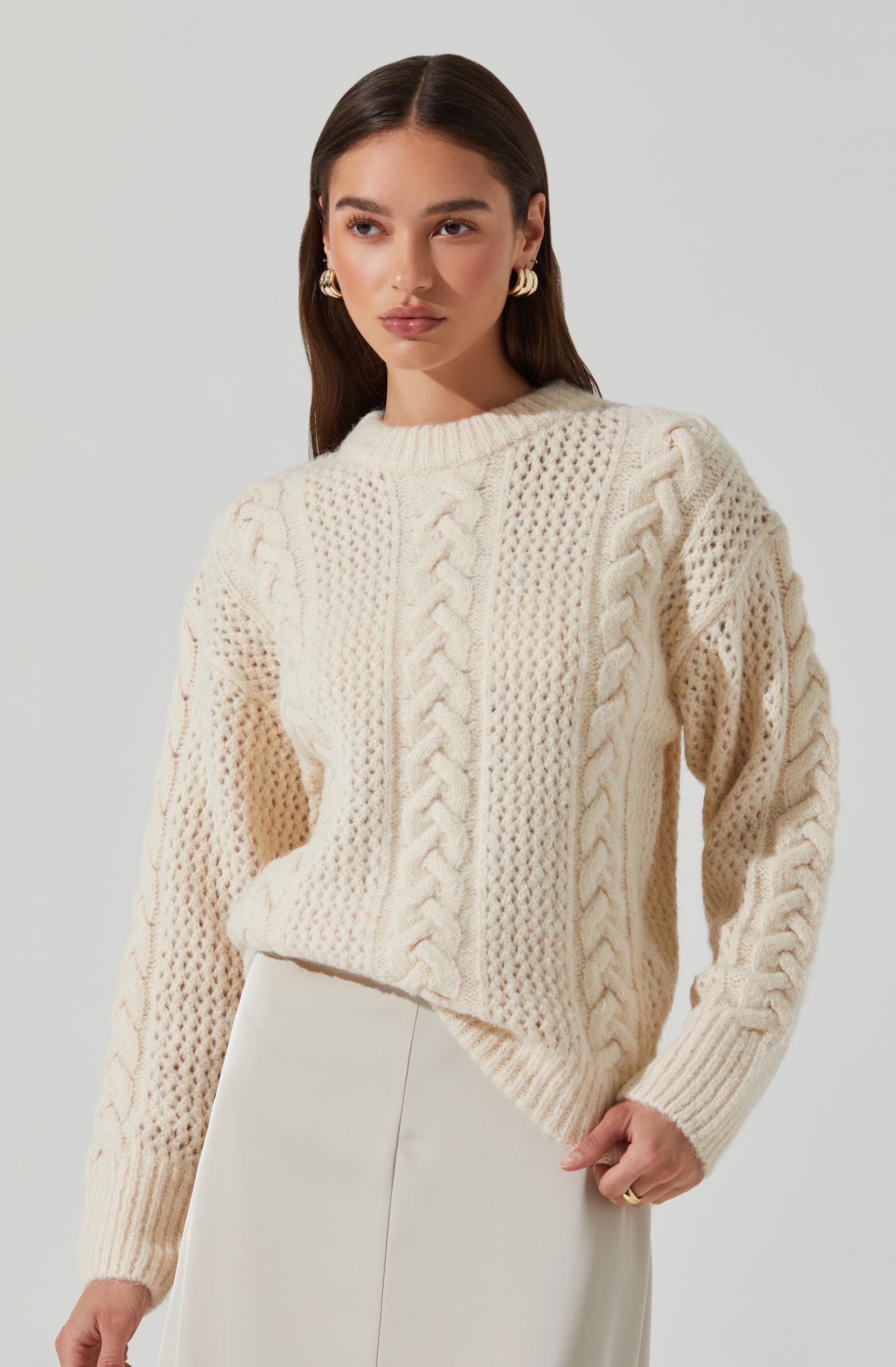 A woman is wearing a cream-colored chunky knit sweater with various cable and textured patterns. The sweater has a round neckline and long sleeves. She is also wearing a pair of elegant, gold hoop earrings with pearl accents.