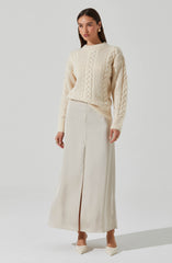A woman is wearing a cream-colored, chunky knit sweater with cable patterns and long sleeves. She pairs it with a high-waisted, floor-length cream skirt that has a front slit. She completes the outfit with pointed-toe heels in a matching cream color.