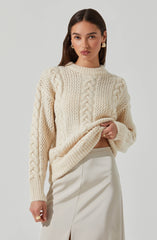 A woman is wearing a cream-colored, chunky knit sweater with various cable knit and textured patterns, and a high crew neckline. She is also dressed in an off-white or light beige satin skirt with a front slit. She accessorizes with large gold hoop earrings and a gold ring on her left hand.