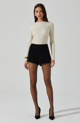 A woman is wearing a cream-colored, long-sleeved, ribbed knit sweater with a round neckline. She is also wearing high-waisted black shorts and sheer black tights. Her outfit is accessorized with gold hoop earrings, a gold bracelet on her right wrist, and a gold ring on her right hand. She has black pointed-toe high heels.