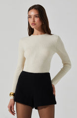 A woman is wearing a fitted, long-sleeved, cream-colored ribbed sweater with a crew neckline. She pairs it with high-waisted, black textured shorts. She accessorizes with a gold bracelet on her left wrist and a ring on her left hand.