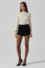 A woman is wearing a cream-colored, long-sleeved, ribbed sweater paired with high-waisted black shorts. She has on sheer black tights and black high-heeled shoes. She accessorizes with a large gold bracelet on her right wrist and small earrings.