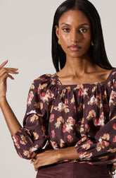 A woman wearing a long-sleeved blouse with a floral pattern in shades of pink, beige, and white on a dark purple background. The blouse has a square neckline with gathered fabric detail. She is also wearing gold earrings.