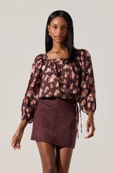 A woman is wearing a dark floral blouse with puffed long sleeves gathered at the wrists, and a square neckline with a slight ruffle detail. She pairs it with a high-waisted, burgundy suede mini skirt that has visible seam details and two small back pockets. She accessorizes with gold earrings and a ring on her right hand.
