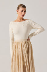 A woman is wearing a cream-colored long-sleeve sweater with small, three-dimensional flower embellishments on the upper chest and shoulders. She pairs it with a high-waisted, beige, pleated skirt made of lightweight, flowy fabric.