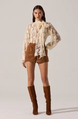 The woman is wearing a long-sleeved, cream-colored lace blouse with a large bow tied at the neck. She has paired it with high-waisted, brown shorts and knee-high brown suede boots.