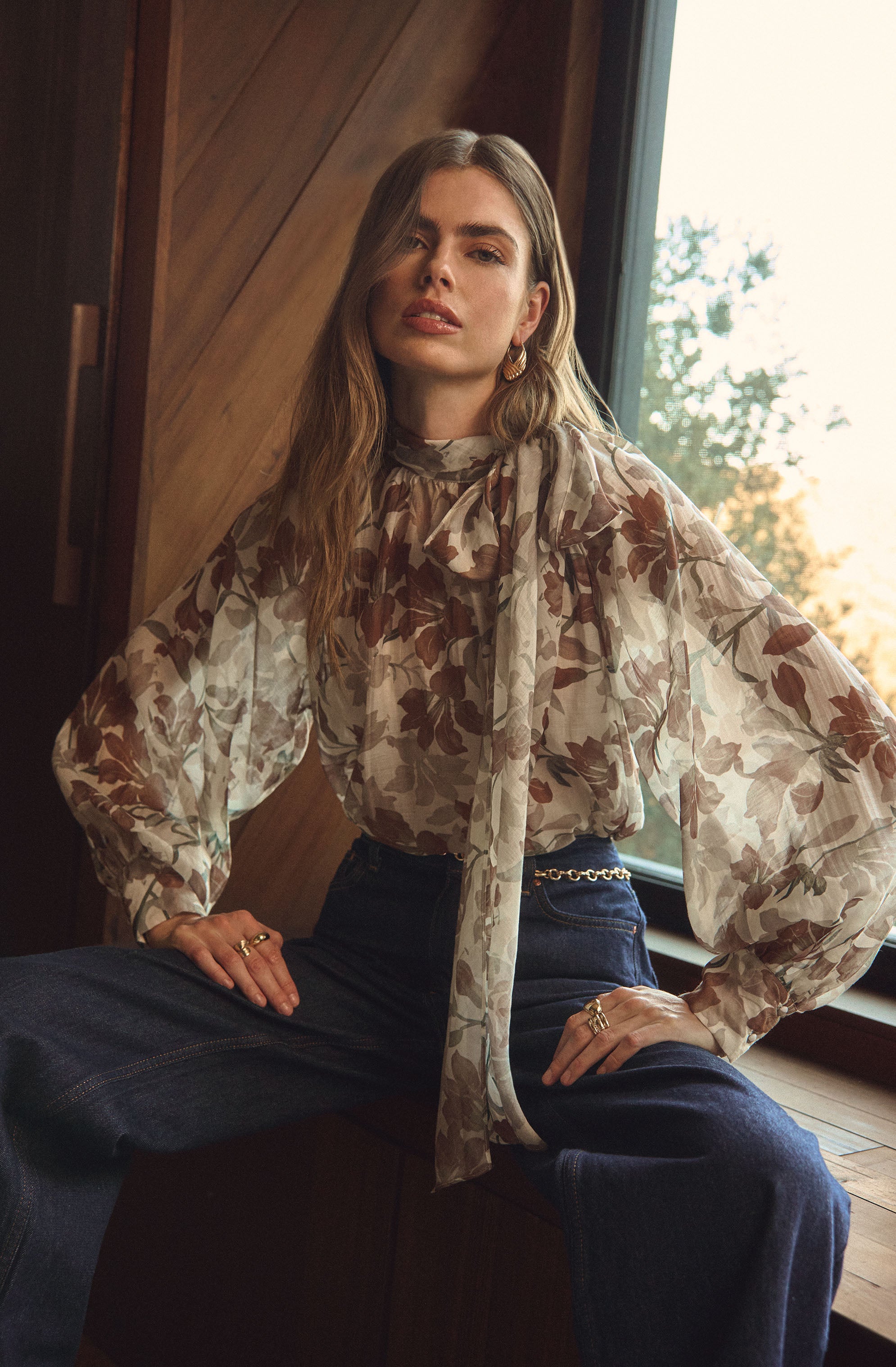 A woman is wearing a long-sleeved blouse with a floral pattern in shades of brown and beige, featuring a high neckline with a bow tie. She has paired it with dark high-waisted jeans that are slightly loose-fitting. She accessorizes with gold earrings, a gold chain belt, and multiple rings.