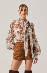 A woman is wearing a long-sleeved blouse with a floral print in shades of brown, beige, and cream, featuring a large bow tie at the neckline. She pairs it with high-waisted, brown shorts made of a suede-like fabric. She accessorizes with large, gold hoop earrings.