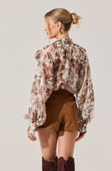 A woman is wearing a sheer, long-sleeved blouse with a floral pattern in shades of pink, beige, and brown, featuring puffed sleeves and a high neckline. She pairs it with high-waisted brown shorts and dark brown knee-high boots. She has her blonde hair styled in a low, messy bun and is accessorized with large gold earrings and a ring on her left hand.