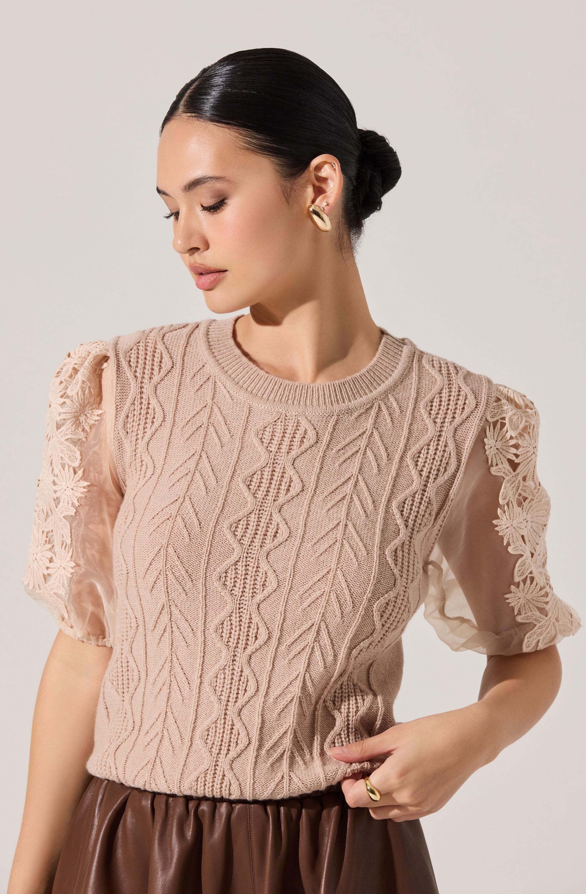 A woman is wearing a beige cable knit sweater with intricate patterns and puffed, lace-embellished short sleeves. She has paired it with a high-waisted, brown leather skirt.
