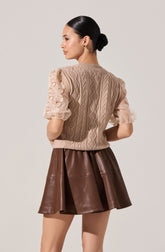 A woman is wearing a beige knitted vest with a cable and floral pattern, layered over sheer, puffed short sleeves with floral lace detailing. She pairs this with a brown leather skirt that has a flared, pleated design.