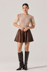 The woman is wearing a beige crochet top with short puff sleeves that have floral cutout patterns. She pairs it with a high-waisted, brown, leather-like A-line mini skirt. She completes the outfit with tall, black, knee-high boots with pointed toes and block heels.