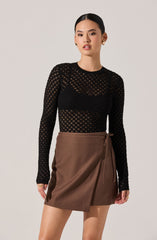 A woman is wearing a black, long-sleeved, see-through, knitted top with a diamond pattern. She has paired it with a brown, wrap-style skirt that ties at the side with a matching fabric belt.
