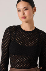 A woman is wearing a black long-sleeved, semi-sheer, knit top with a diamond-shaped openwork pattern. The top has a round neckline and features a solid black horizontal band across the chest. She has paired it with light brown high-waisted pants that have a bow tied at the waist. She is accessorized with gold hoop earrings and has her dark hair styled in a sleek, pulled-back look.