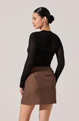 A woman is wearing a black, long-sleeved, open-knit top paired with a high-waisted brown skirt that falls above the knees. The top has a see-through pattern, and she has her dark hair styled in a sleek bun. She is accessorized with gold hoop earrings.