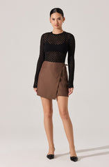 A woman is wearing a black, long-sleeved, see-through knit top with a geometric pattern. She pairs it with a brown wrap-style mini skirt that ties on the side. She completes the outfit with black pointed-toe heels.