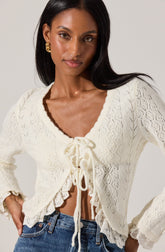 A woman is wearing a cream-colored knitted cardigan with a lace trim along the edges and a front tie closure. The cardigan features a textured, open-knit pattern and ruffled details at the hem and cuffs.
