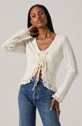 A woman is wearing a cream-colored, long-sleeved, knit cardigan with a delicate, open lace pattern. The cardigan features a V-neckline with a drawstring tie at the front and ruffled edges along the hem and cuffs. She pairs it with high-waisted blue jeans.