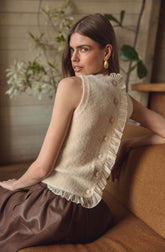 A woman is wearing a sleeveless, cream-colored top made of a fuzzy knit fabric. The top features a row of decorative buttons along one side, with ruffled trim alongside the buttons. The top appears to have a slightly ruffled hem.