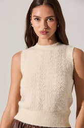 A young woman with long brown hair is wearing a sleeveless, cream-colored knit sweater with a textured, cable pattern design. She has a neutral facial expression, with minimal makeup and small earrings. The background is plain and light-colored.