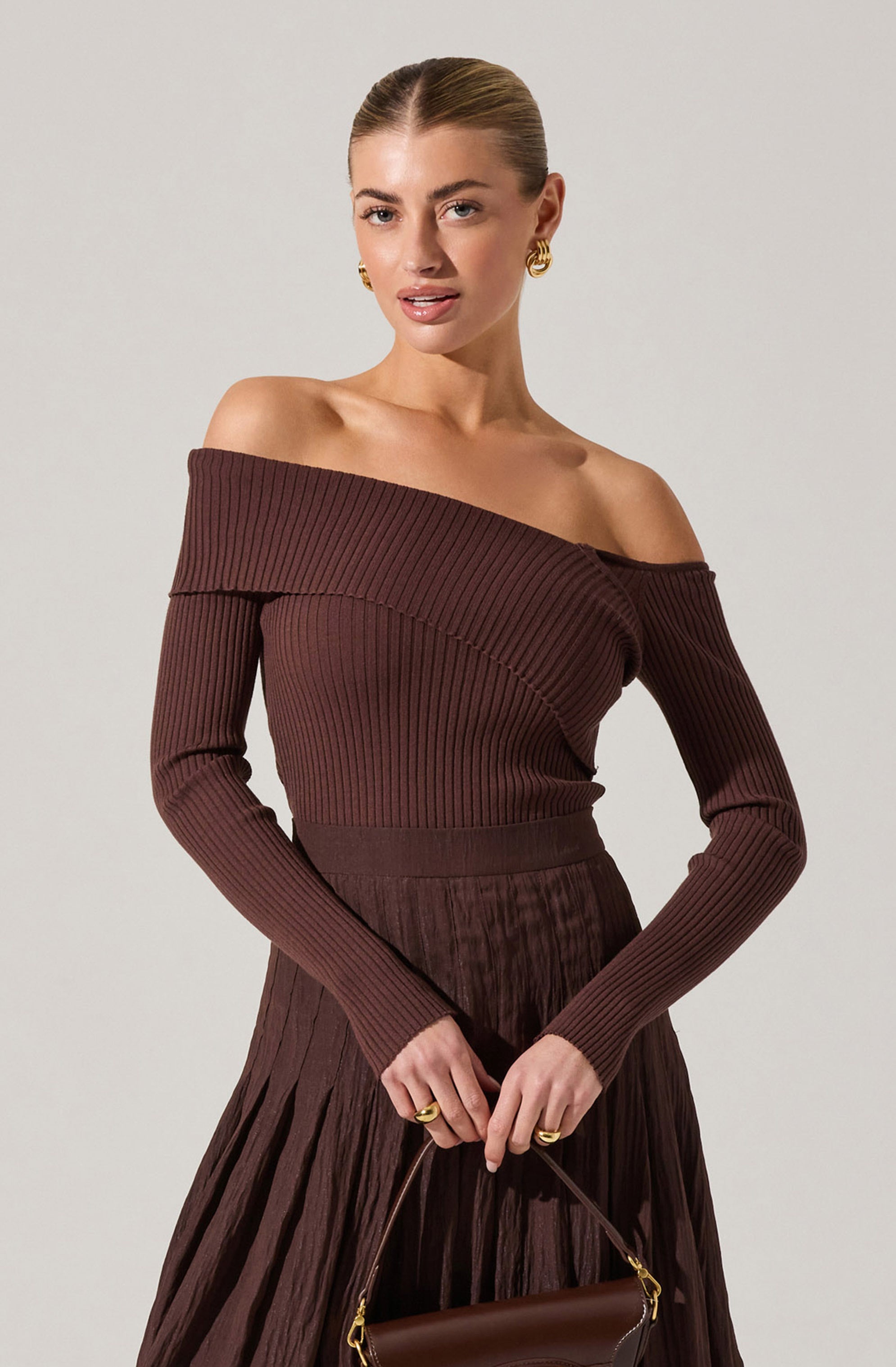 A woman is wearing a fitted, long-sleeved, off-the-shoulder ribbed sweater in a deep brown color. The sweater features a wide neckline that drapes over her shoulders, exposing her collarbones. She pairs it with a matching high-waisted, pleated skirt in a similar brown shade. She accessorizes with gold hoop earrings and rings, and holds a small brown handbag with a gold clasp and handle.