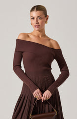 A woman is wearing a fitted, long-sleeved, off-the-shoulder ribbed sweater in a deep brown color. The sweater features a wide neckline that drapes over her shoulders, exposing her collarbones. She pairs it with a matching high-waisted, pleated skirt in a similar brown shade. She accessorizes with gold hoop earrings and rings, and holds a small brown handbag with a gold clasp and handle.