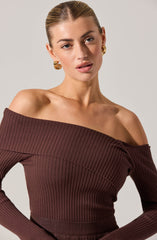 A woman is wearing an off-the-shoulder, long-sleeved ribbed top in a deep brown color. The top features a wide neckline that sits below the shoulders, exposing her collarbones. She is accessorized with large, gold hoop earrings.