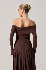 A woman is wearing a coordinating set in a deep brown color. The off-the-shoulder top has long sleeves and features a wide, folded-over neckline with a ribbed texture. The high-waisted skirt is also ribbed at the waist and transitions into pleated fabric that extends to the floor. She accessorizes with gold earrings and a ring, and her hair is styled in a sleek bun.