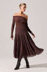 The woman is wearing an off-the-shoulder, long-sleeved, ribbed knit top in a deep brown color. She is also dressed in a matching brown, pleated, mid-length skirt with a textured fabric that creates a flowy effect. Her outfit is complemented by black knee-high boots.