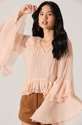 A woman is wearing a light peach-colored blouse with a V-neckline. The blouse features vertical pleats on the front, ruffled details along the hem and the wide, billowy sleeves, which have layered ruffles at the cuffs. She is also wearing brown shorts, and has long, dark, wavy hair. She is accessorizing with colorful, dangling earrings.