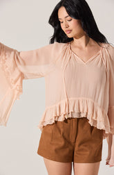 A woman is wearing a light peach, loose-fitting blouse with a V-neckline, ruffled details along the hem and sleeves, and a soft, flowing fabric. She pairs it with high-waisted brown shorts that have a relaxed fit and an elastic waistband.
