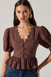 The woman is wearing a brown textured blouse with a scalloped neckline, short puff sleeves, and a peplum hem. The blouse has a row of fabric-covered buttons down the front. She is accessorized with large, gold hoop earrings.