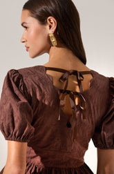A woman is wearing a brown, textured dress with puffed short sleeves. The dress features a cut-out back with a bow-tie closure made of matching fabric ribbons. She has large, ornate gold earrings.