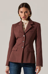 A woman is wearing a tailored, dark brown blazer with a structured fit, notch lapels, and three visible buttons on the front. She has on a high-neck, lace blouse underneath that is cream-colored with delicate floral patterns. She pairs the blazer with dark blue jeans and accessorizes with large, gold hoop earrings and a gold ring on her right hand.