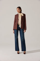 A woman is wearing a tailored, dark brown blazer with notch lapels and a button closure. Underneath, she has a cream-colored lace blouse with ruffled detailing around the neckline and shoulders. She pairs this with high-waisted, straight-leg blue jeans and dark brown high-heeled shoes. She accessorizes with gold hoop earrings and a gold ring.