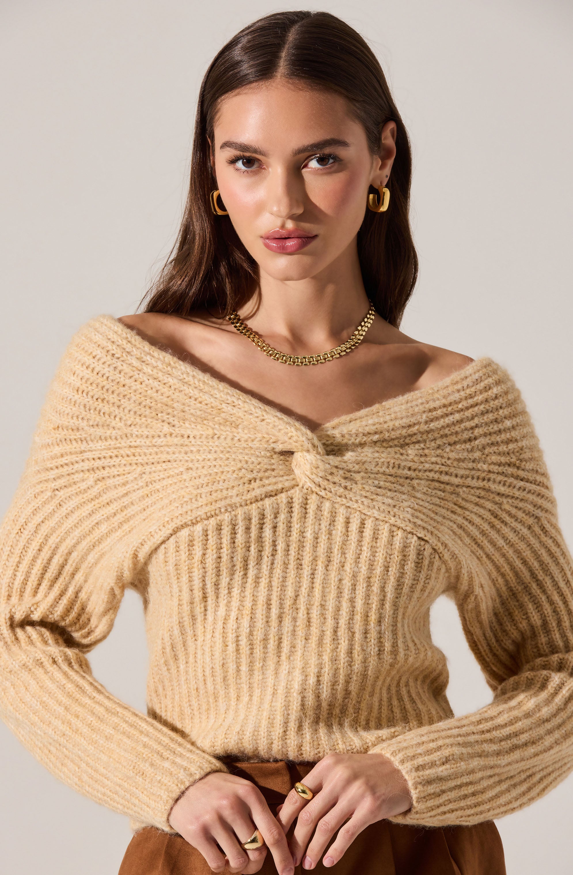 A young woman is wearing a beige, chunky knit sweater with a twisted front neckline. She accessorizes with gold hoop earrings, a gold chain necklace, and rings on her fingers.