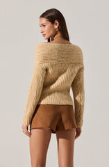 A woman is wearing a beige, chunky, ribbed knit sweater with a wide, folded collar that extends over her shoulders. She pairs it with high-waisted brown suede shorts. She accessorizes with gold hoop earrings, a gold chain necklace, and a jewelry ring on her left hand.