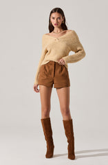 A woman is wearing a chunky, off-the-shoulder beige knit sweater with a wide neckline and long sleeves. She pairs it with high-waisted brown shorts that have a pleated front. She completes the look with tall, brown suede over-the-knee boots.