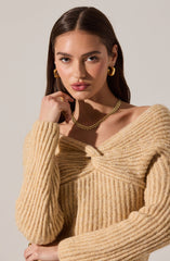 A young woman is wearing a beige, chunky ribbed knit sweater with a twist detail at the neckline. She accessorizes with large gold hoop earrings, a gold chain necklace, and a gold ring on her right hand.