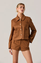 A woman is wearing a matching brown suede jacket and shorts set. The jacket has a rounded collar, four large buttons down the front, and two front pockets. The shorts are high-waisted and have a tailored fit.