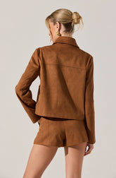 A woman is wearing a brown suede jacket and matching brown suede shorts. The jacket has a collar and long sleeves, and the shorts are fitted with a simple waistband. The outfit appears stylish and coordinated in tone and material.