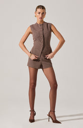 A woman is wearing a sleeveless, button-up vest and matching shorts in a small checkered pattern. The vest has a stand-up collar, is fitted at the waist, and features decorative buttons down the front. She pairs the outfit with sheer brown tights and pointed-toe high-heeled shoes. She accessorizes with gold earrings and a gold bracelet.