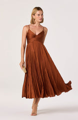A woman is wearing a rust-colored, pleated dress with a fitted, ruched bodice and thin spaghetti straps. She is holding a small, rectangular clutch and is wearing open-toed, strappy heels. She has shoulder-length blonde hair and is accessorized with large, square earrings.