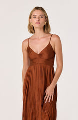 The woman is wearing a brown, pleated dress with thin spaghetti straps, featuring a gathered detail at the bust and a flowing, textured skirt.