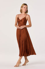 A woman is wearing a rust-colored, pleated, midi-length dress with spaghetti straps. The dress has a fitted bodice with ruching details, flowing pleats from the waist down, and a v-neckline. She is also wearing beige high-heeled sandals.