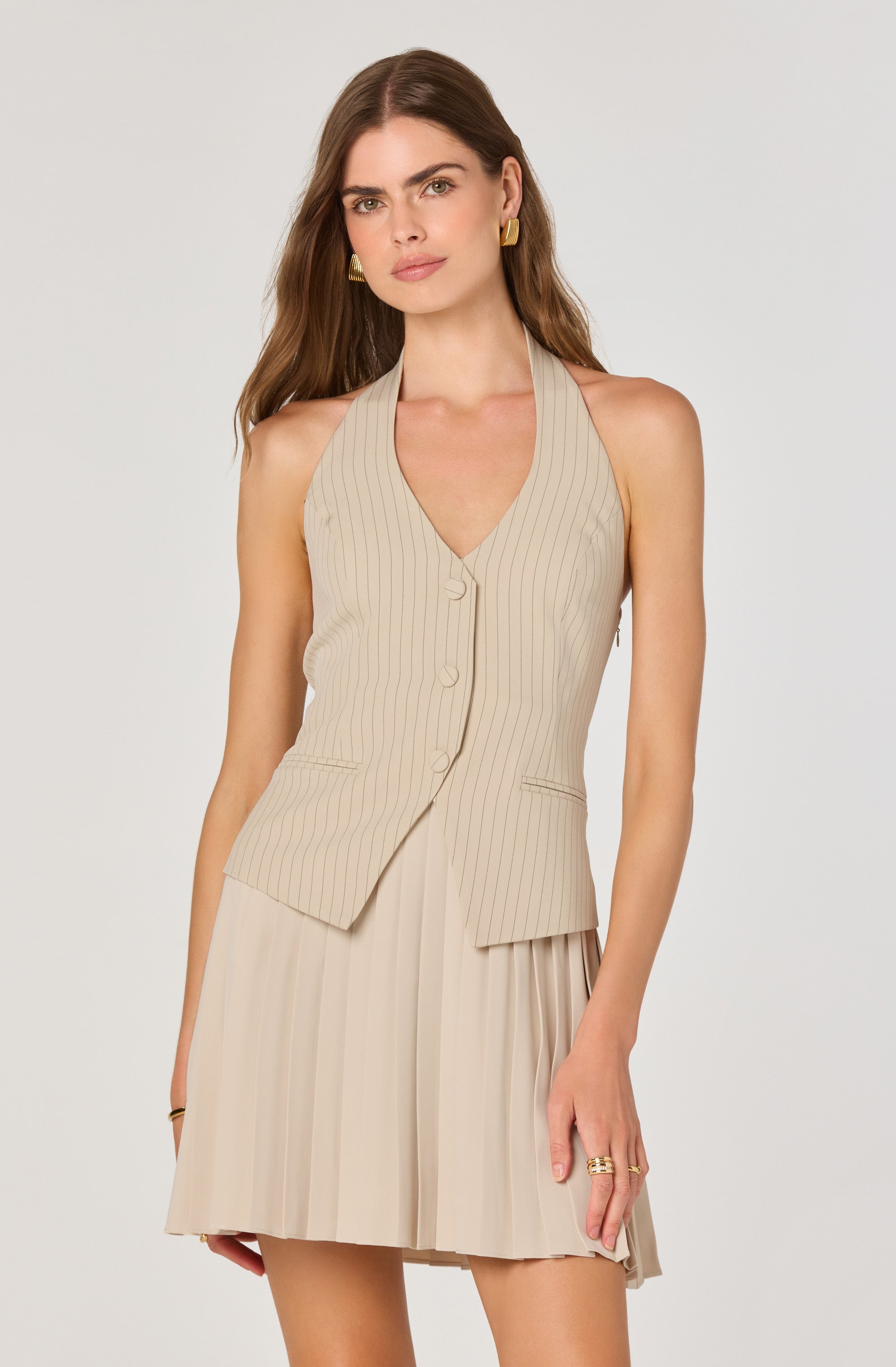 A woman is wearing a sleeveless beige vest with thin vertical stripes and a V-neckline, featuring buttons down the front and two small pockets at the waist. She pairs it with a matching pleated beige skirt. She accessorizes with gold earrings, rings, and a bracelet.