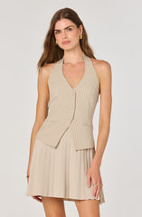 A woman is wearing a sleeveless beige vest with thin vertical stripes and a V-neckline, featuring buttons down the front and two small pockets at the waist. She pairs it with a matching pleated beige skirt. She accessorizes with gold earrings, rings, and a bracelet.