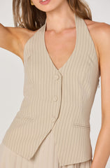 A woman is wearing a sleeveless, beige pinstriped vest with vertical black stripes. The vest features a V-neckline, a front button closure with three buttons, and two small slit pockets on either side. The fabric appears lightweight and tailored for a chic, professional look. The woman is also wearing large, gold rectangular earrings.